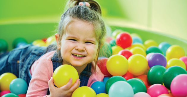 Smiling toddler playing in a colourful ball pit at an indoor kids’ play area, holding a yellow ball