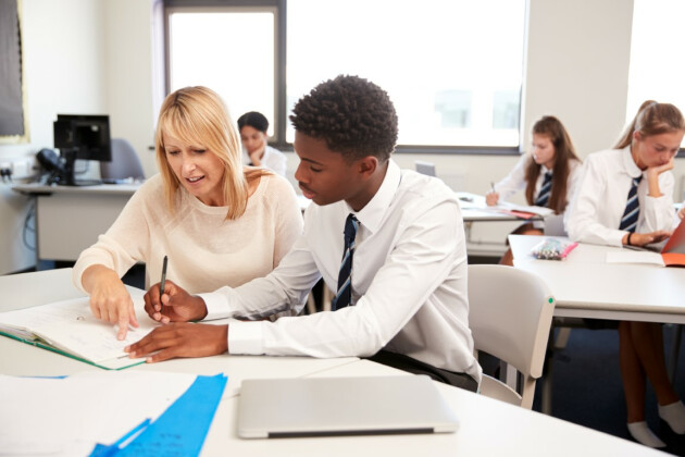 A young male student is being helped by his teacher, their sat at a table together looking at a workbook