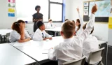 A classroom of students, the view is behind a boy's head with his arm raised for his teacher