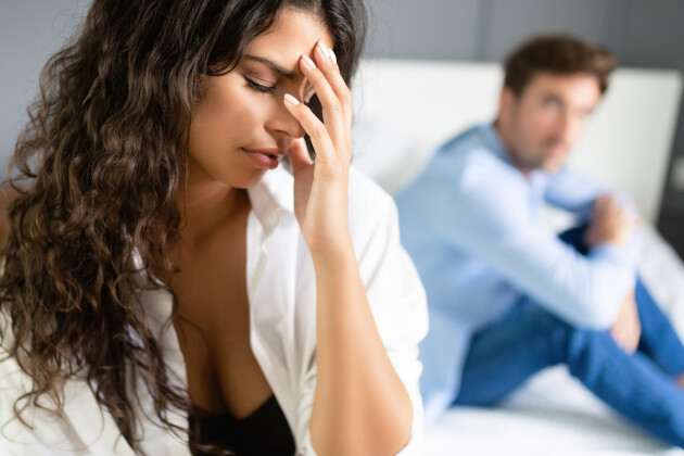 A woman holds her head in her hands sat on the bed with her husband in the background