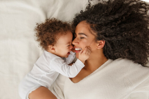 A mother and her baby smile at each other while laid atop a bed, the baby is holding her face