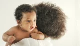 Mother with natural curly hair holding smiling baby wrapped in white muslin cloth in bright studio portrait photo