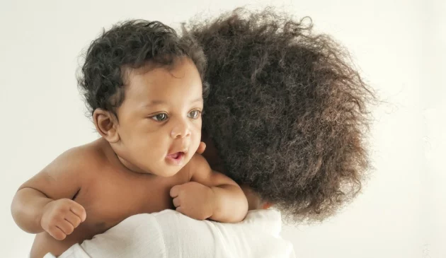 Mother with natural curly hair holding smiling baby wrapped in white muslin cloth in bright studio portrait photo