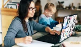A mother working on a laptop with her child next to her drawing
