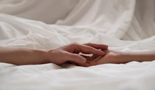 A couple's hands reaching for each other over white bedsheets