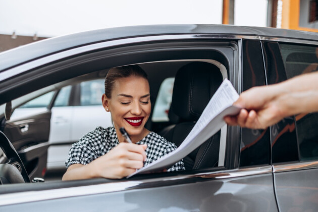 A woman smiling signs a car contract out of the window while sat in the car