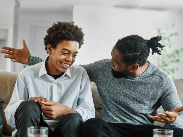 A father and son talking while sat on a sofa, the son is smiling while the dad embraces him