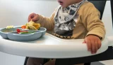 Toddler in dinosaur bib self-feeding vegetables and mashed food from divided plate in high chair at mealtime at home