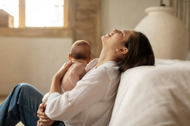 A woman holds her baby but appears to be stressed with her head back against a bed