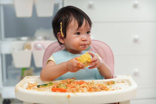 Happy baby in pink high chair enjoying baby-led weaning with corn on the cob and colorful vegetables spread on feeding tray