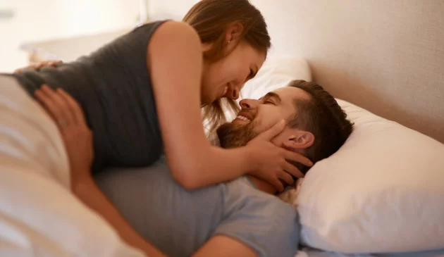A happy couple laid in bed, the woman is on top of the man under the duvet cover, both are clothed