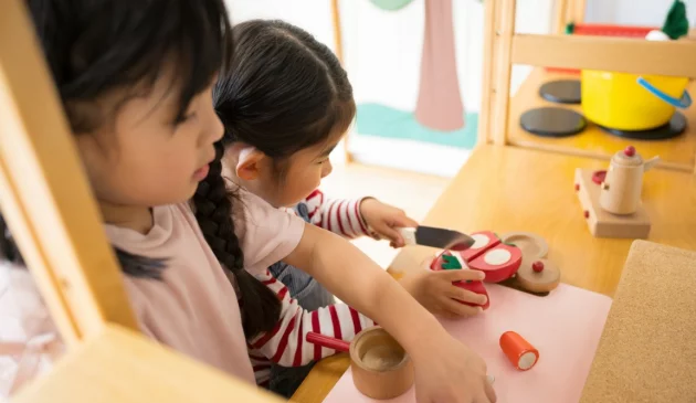 wo young girls playing with wooden toy kitchen set and pretend food at table in bright nursery playroom setting