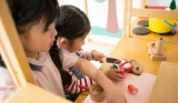 wo young girls playing with wooden toy kitchen set and pretend food at table in bright nursery playroom setting