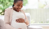 Pregnant woman sitting on couch smiling while gently holding baby bump during third trimester at home