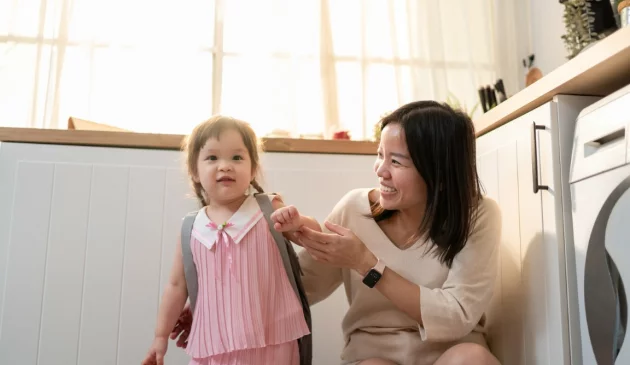 Mother helping young daughter with pink backpack prepare for nursery in bright modern kitchen with natural sunlight