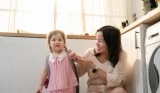 Mother helping young daughter with pink backpack prepare for nursery in bright modern kitchen with natural sunlight