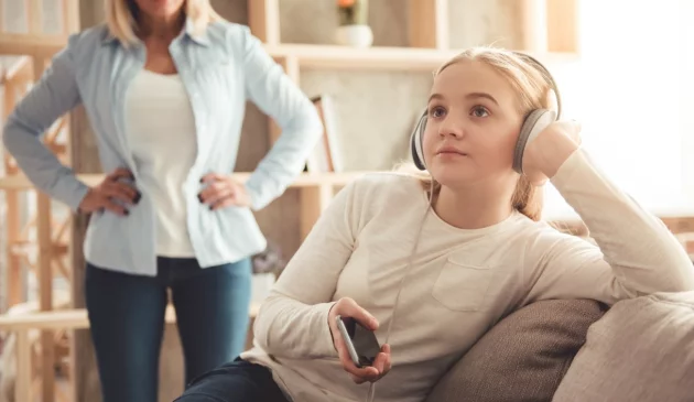 A teenager daughter with headphones on ignores her mother in the background