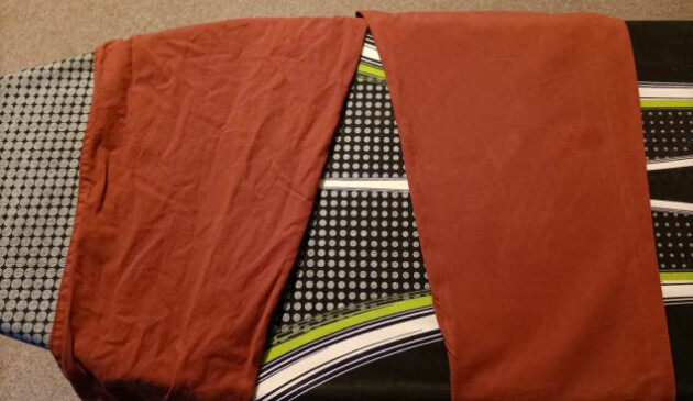 A pair of terracotta-coloured trousers laid out on a black, grey, white and lime green ironing board. One trouser leg is wrinkled and creased, the other is smooth.