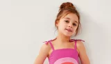A little girl wearing a pink Monsoon dress smiles at the camera against a white wall