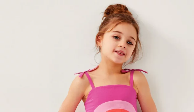 A little girl wearing a pink Monsoon dress smiles at the camera against a white wall