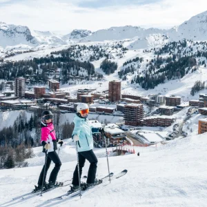 Shot of two young skiers smiling on piste with resort, ski lifts and mountains of La Plagne in background. Photo credit: Brendan Le Peru