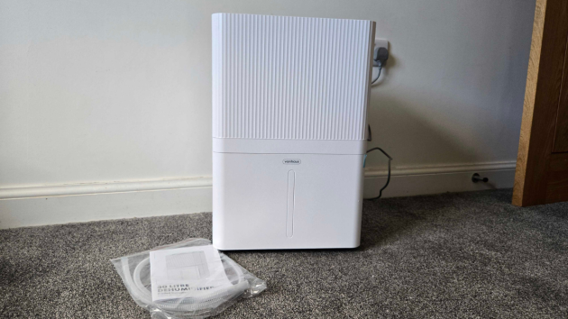 The front of a white VonHaus dehumidifier with a ribbed upper panel and a vertical water-level window on the lower tank, with a bagged hose and manual on the carpet in front