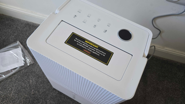 Top panel of the dehumidifier with a black and yellow “Important!” sticker warning that the unit must stand upright for 24 hours before operation, next to a blank round display and a row of touch buttons