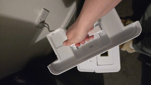 A hand pulling the white water tank out of the dehumidifier in a dimly lit room, with the unit’s top panel visible below