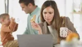 Woman works and eats toast while husband attends to baby in blurred background